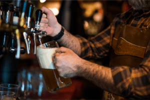 A male bartender is pouring frothy tap beer into a thick glass mug.