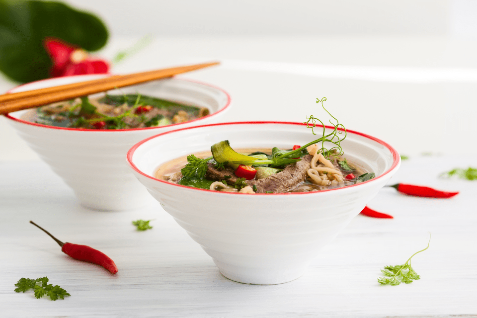 Two white bowls containing Asian-style meat and noodles accompanied by some chopsticks and red peppers.