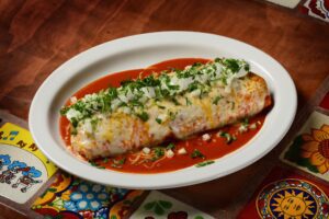 A burrito on a white plate covered in red sauce, white cheese, and green herbs.