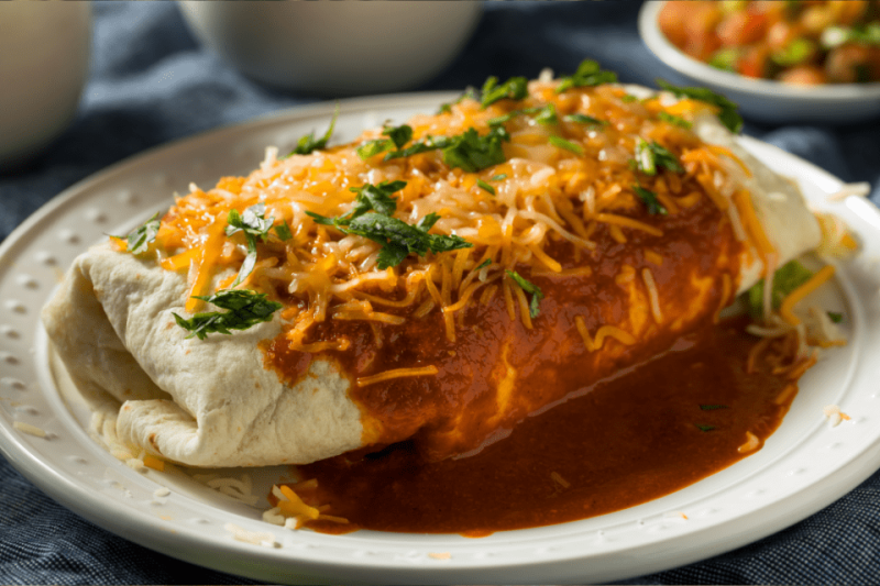 A burrito smothered in red sauce and yellow cheese topped with cilantro.