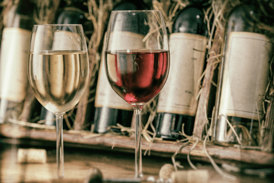 A glass of white wine and a glass of red wine in front of beautiful bottles of wine.