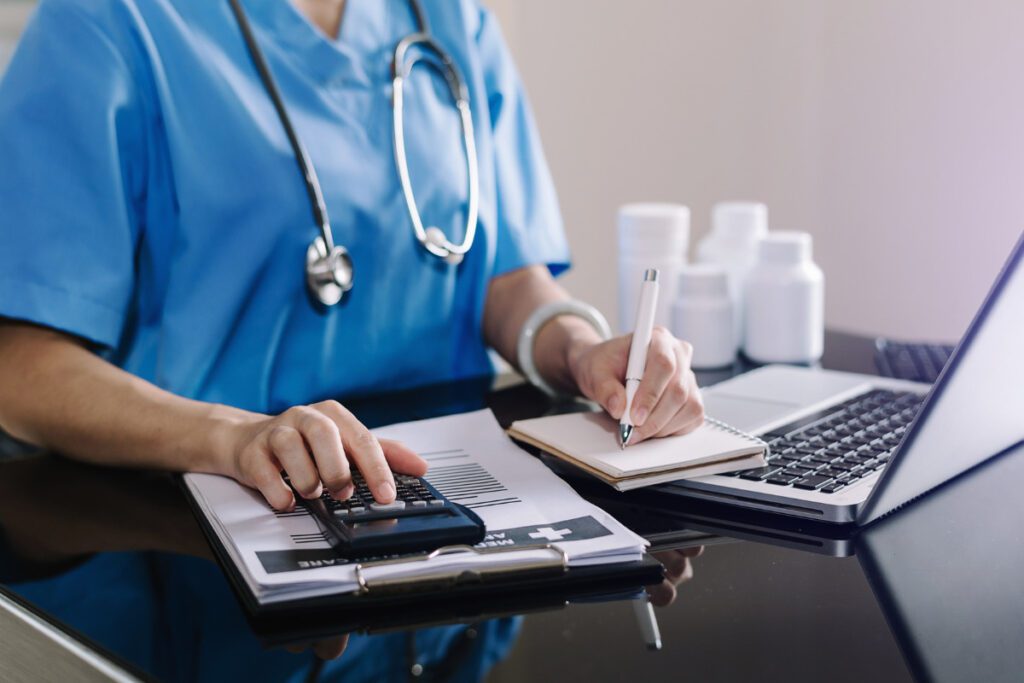 Photo of healthcare worker in front of computer, using a calculator to add up medical costs