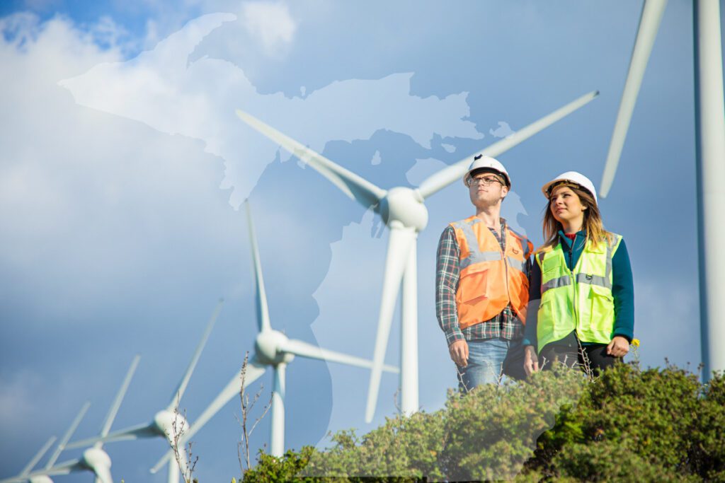 Climate Change - Photo of two workers wearing safety vests and hard hat standing on hill in front if wind turbines.