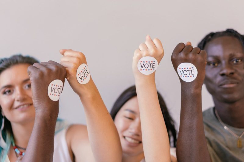 3 young people smiling holding their arm up with stickers on hand reading "Vote"