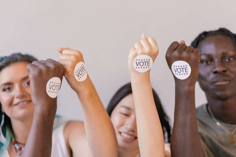 3 young people smiling holding their arm up with stickers on hand reading "Vote"