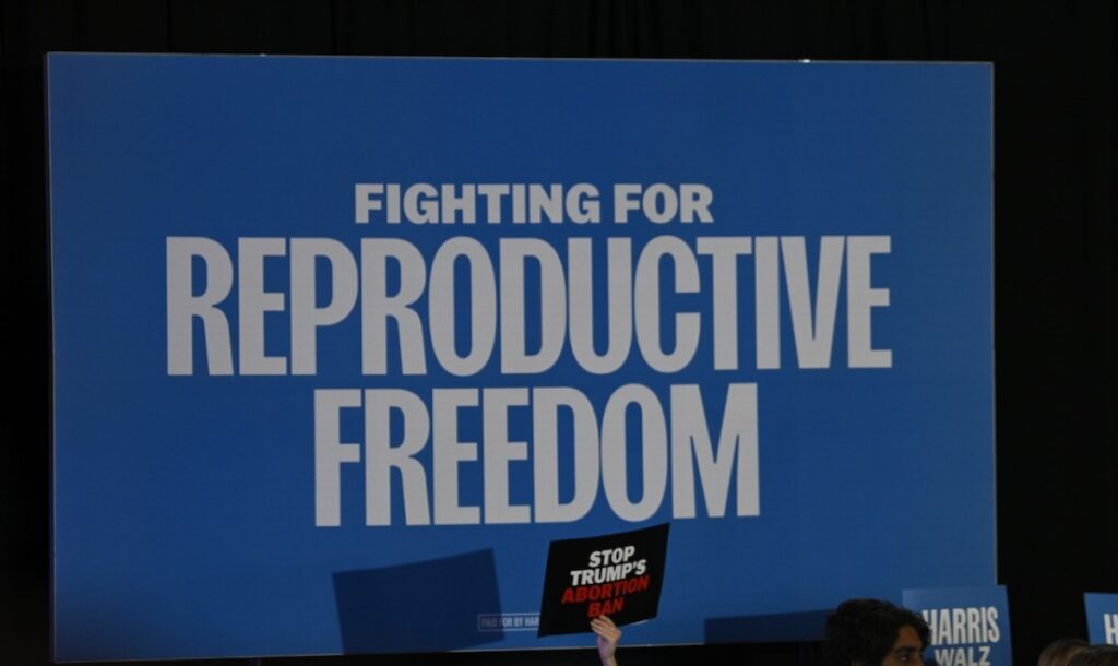 Picture of Hands holding a sign that reads "Stop Trump's Abortion Ban" in front of larger sign reading "Fighting for reproductive freedom"