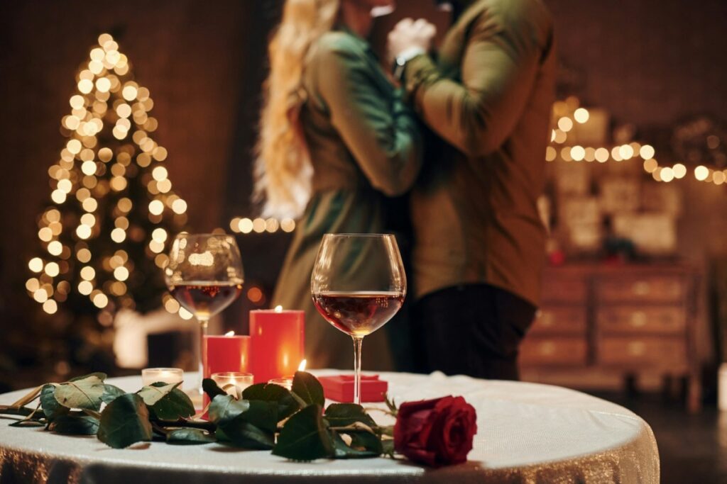 Two wine glasses in the foreground with a couple holding hands and standing in the background against a Christmas backdrop.