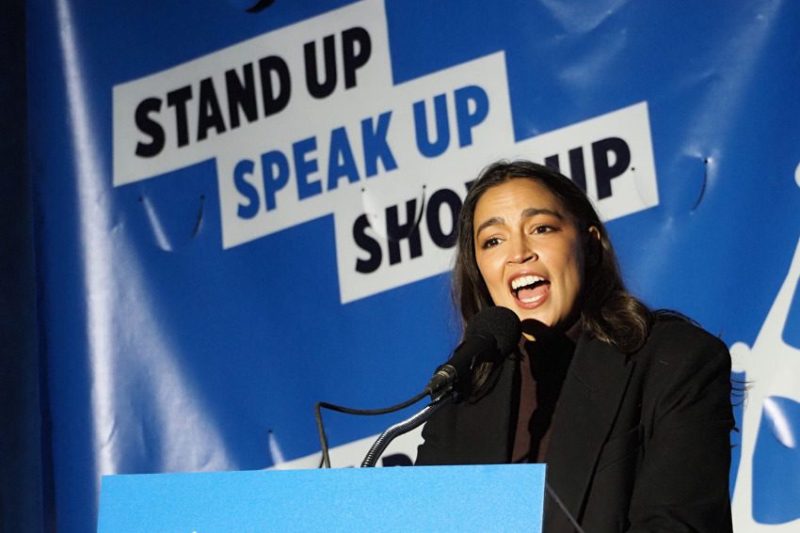 AOC speaking at an event in front of blue background.