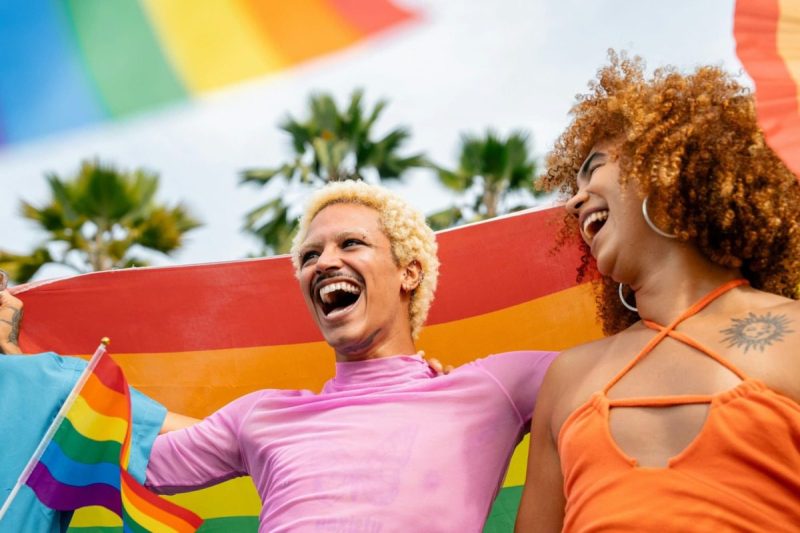 two people smile in front of a pride flag