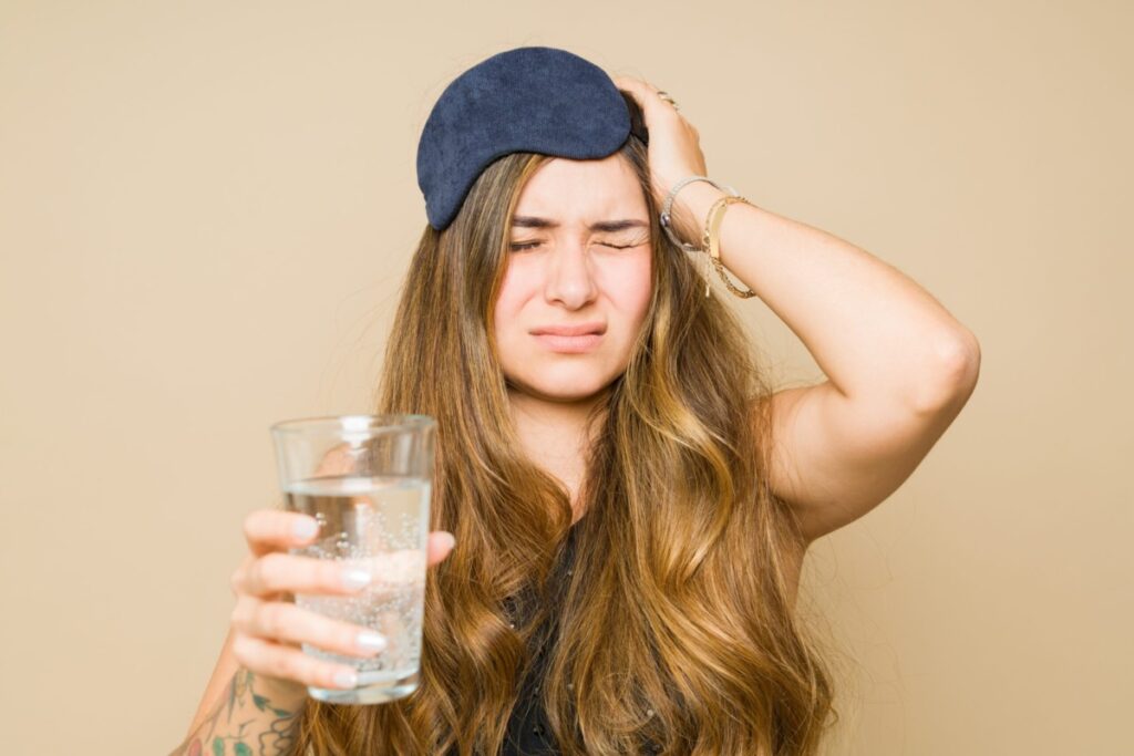 a hungover woman grimaces while holding a glass of water and her head