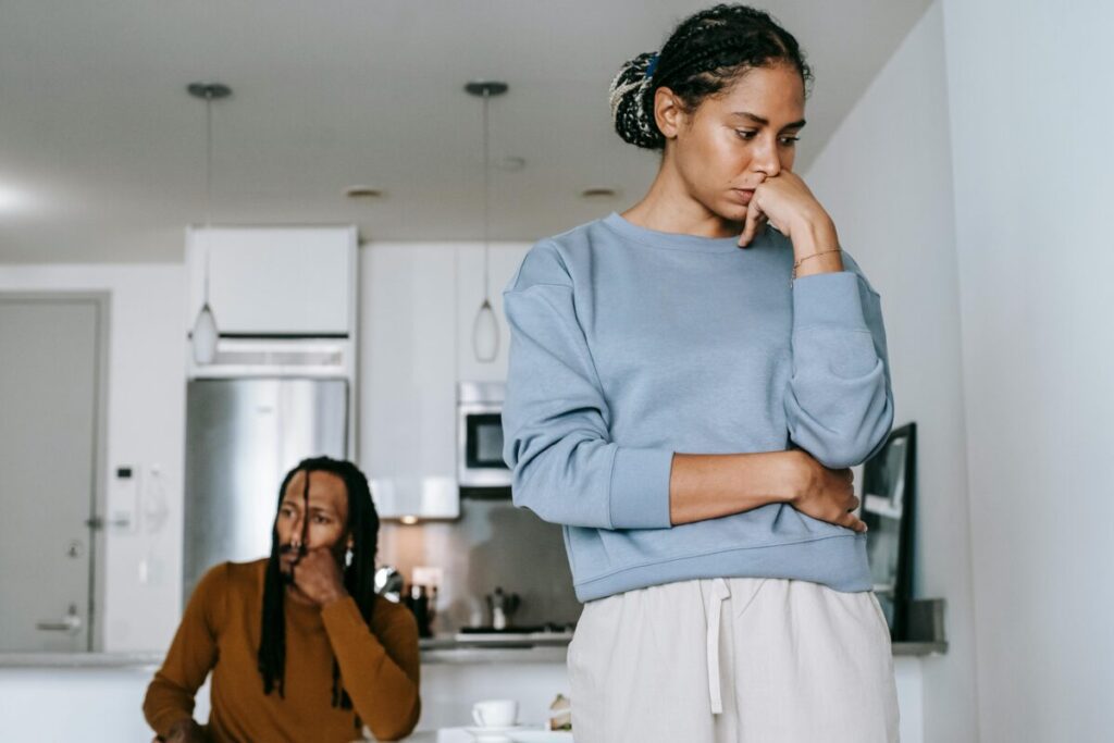 Photo of couple. One is standing facing the camera, the other in the background on a couch. Both looking worried.