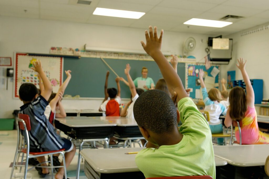funding and investing in public schools. Kids in a classroom responding to the teacher's question by raising their hands.