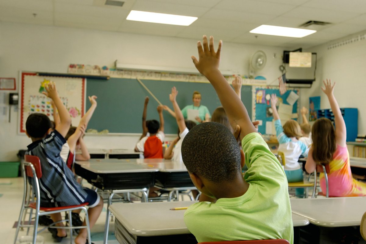 funding and investing in public schools. Kids in a classroom responding to the teacher's question by raising their hands.